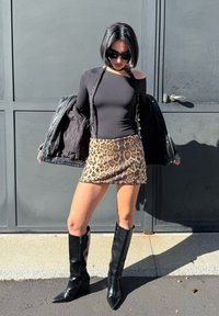 Crna bluzica dugih rukava, mini suknja s leopard uzorkom i crne čizmice do koljena s oštrim vrhom. Model drži crnu jaknu. Glatka tekstura i uski kroj.