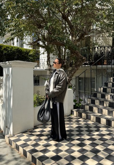 Mujer con gafas de sol, chaqueta de cuero y pantalones negros de Adidas sostiene un bolso tejido en un porche con baldosas a cuadros cerca de las escaleras y un árbol.