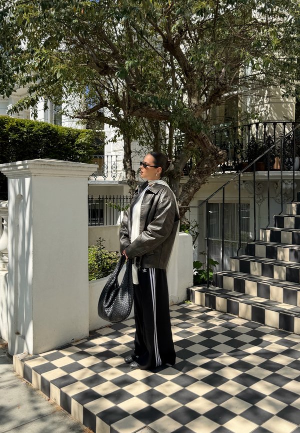 Femme portant des lunettes de soleil, une veste en cuir et un pantalon Adidas noir tient un sac tissé sur un porche en carreaux près des escaliers et d’un arbre.