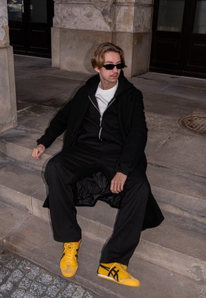 Sudadera con capucha negra con cremallera sobre una camiseta blanca, combinada con pantalones negros holgados y zapatillas amarillas con rayas negras. Escenario urbano minimalista.