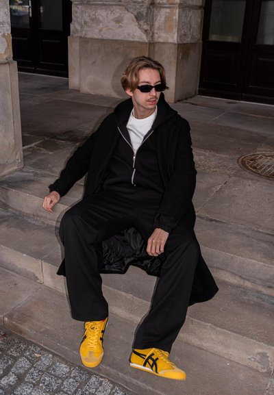 Sudadera con capucha negra con cremallera sobre una camiseta blanca, combinada con pantalones negros holgados y zapatillas amarillas con rayas negras. Escenario urbano minimalista.