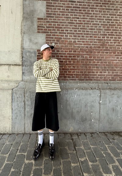 Camisa de manga larga a rayas en verde claro y blanco, pantalones cortos anchos negros, zapatos de vestir negros y calcetines blancos con rayas negras, frente a una pared de ladrillo.