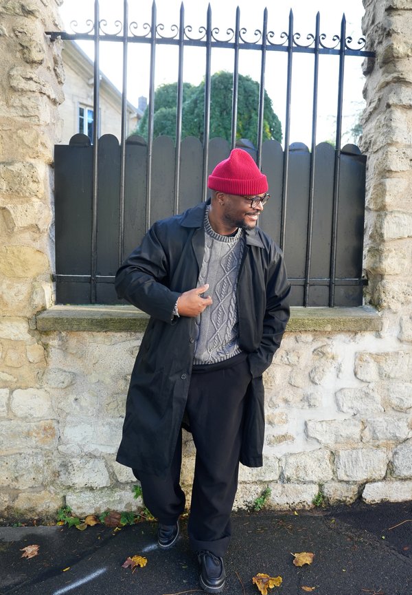 Un homme souriant portant un bonnet en tricot rouge, des lunettes et un manteau foncé se tient près d’un mur en pierre et d’une grille en fer.