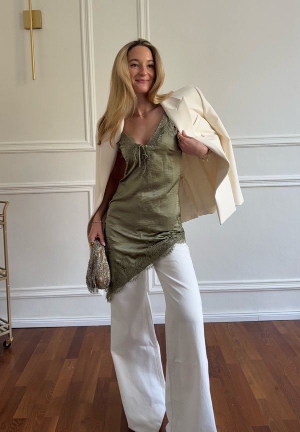 Sieviete zaļā mežģīņu apmalēta slipkleitā pār baltām biksēm, plecos kaila bēša žakete un rokās rokassomiņa ar bārkstīm.