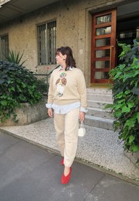 Beżowy sweter z grafiką misia, jasnoniebieska koszula z guzikami, beżowe spodnie, czerwone baleriny i mała biała torebka, na tle teksturowanej ściany.