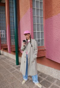 Bežna trench coat s gumbi, svetlo roza kapica, roza srajca, modri džins in bež čevlji. Stoji ob teksturirani roza in oranžni steni.
