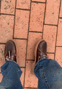 Personne portant des bottes en cuir marron et un jean bleu debout sur un pavement en briques rouges avec des interstices visibles et de petits graviers.