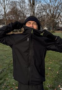 Crna jakna s patentnim zatvaračem i visokim ovratnikom, lagane tkanine i patentni zatvarač sprijeda, nošena s crnim rukavicama na pozadini parka.