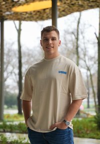 Beige t-shirt med kort ärm och "SPRING" i blått tryck på vänster bröst, matchad med blå jeans. Slät textur och avslappnad passform.