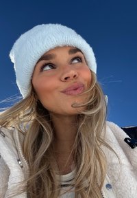 Jeune femme portant un bonnet d'hiver blanc et une veste, regardant vers le haut avec une expression malicieuse sous un ciel bleu clair.