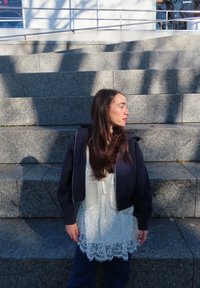 Mujer de cabello largo y oscuro que lleva una chaqueta negra sobre una blusa blanca de encaje, de pie en unos escalones de piedra y mirando hacia su derecha.