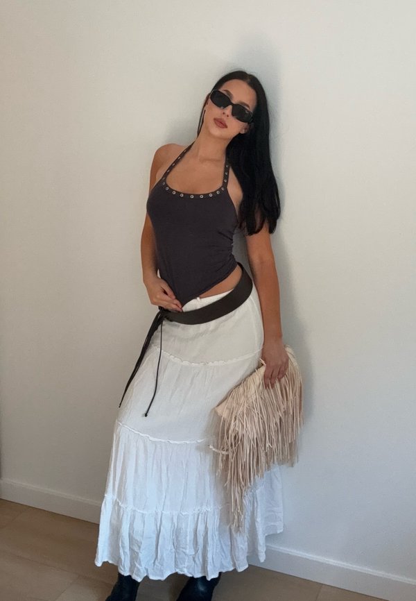 Donna con top nero a collo alto, gonna bianca a balze, cintura nera, occhiali da sole neri e borsa beige con frange in mano, in piedi contro una parete liscia.