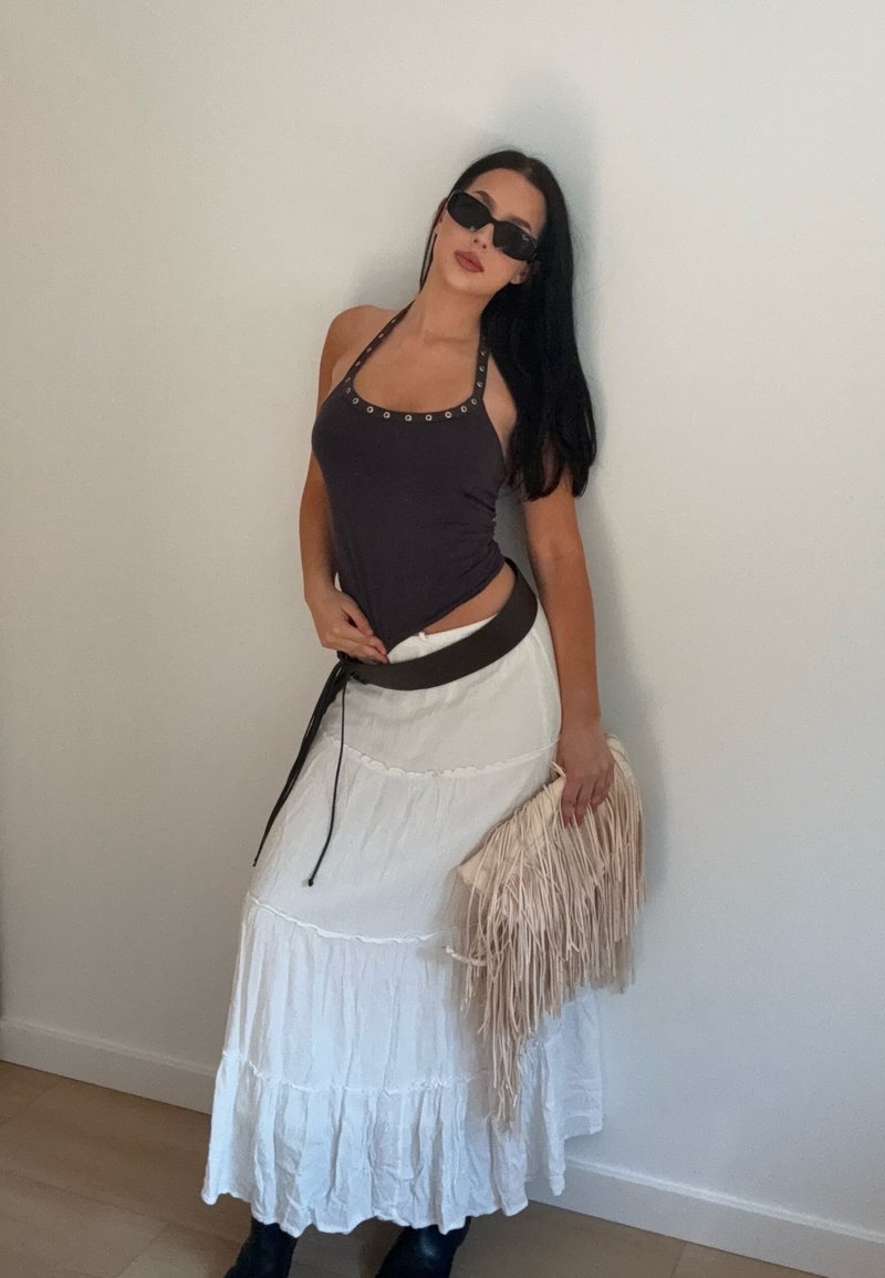 Femme portant un haut à licou noir, une jupe blanche à volants, une ceinture noire, des lunettes de soleil noires, et tenant une pochette beige à franges, debout contre un mur uni.