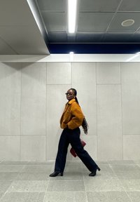 Veste en fourrure marron, pantalon large sombre et bottes à talons, marchant dans un couloir avec des murs gris clair et des lumières au plafond.