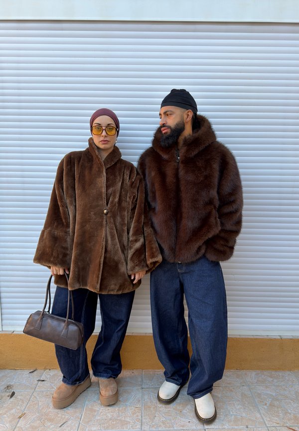 Cappotti in pelliccia sintetica marrone con maniche larghe, abbinati a jeans in denim scuro. La donna tiene una piccola borsa marrone. Lo sfondo presenta una porta avvolgibile bianca.