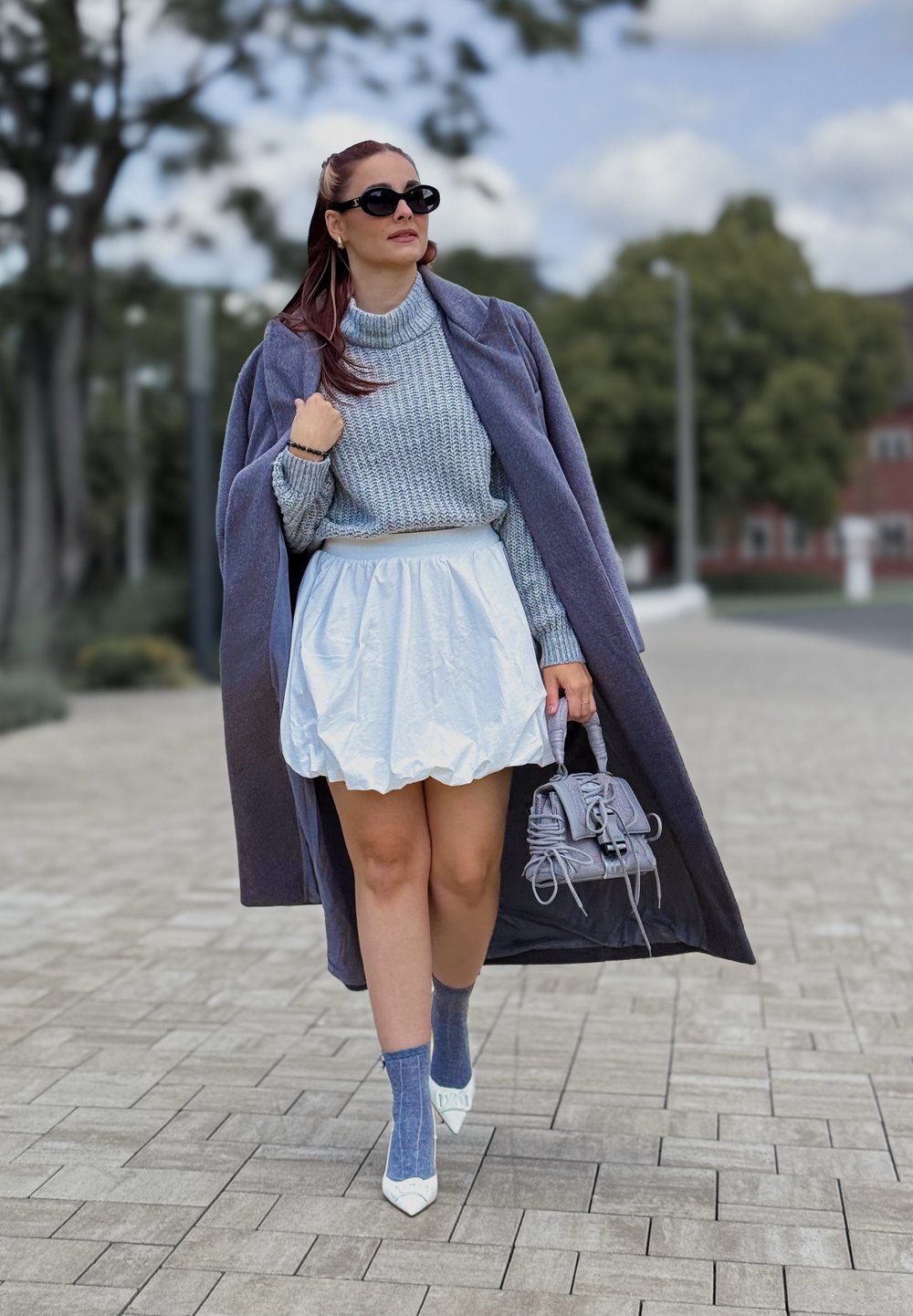 Palton lung gri, pulover tricotat albastru, fustă puff albă, geantă gri, șosete cu model albastre, pantofi albi cu vârful ascuțit, peisaj exterior cu copaci.