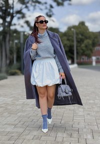 Palton lung gri, pulover tricotat albastru, fustă puff albă, geantă gri, șosete cu model albastre, pantofi albi cu vârful ascuțit, peisaj exterior cu copaci.