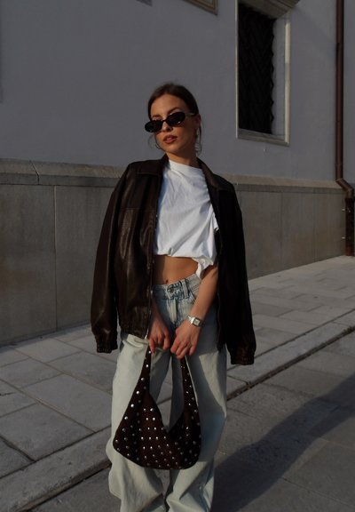 Joven mujer con gafas de sol, top corto blanco, chaqueta de cuero marrón y vaqueros claros, sosteniendo un bolso marrón con tachuelas en una acera de la ciudad.