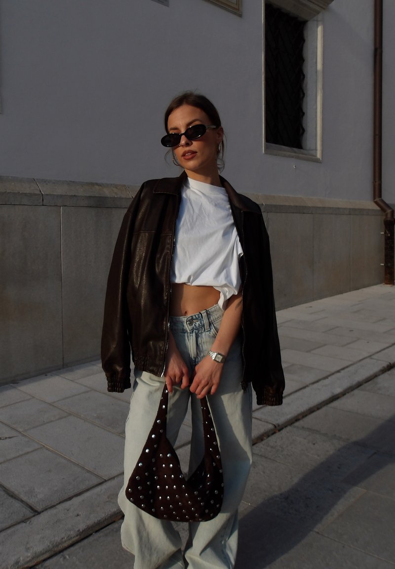 Jeune femme portant des lunettes de soleil, un top court blanc, une veste en cuir marron et un jean clair, tenant un sac à main marron clouté sur un trottoir en ville.