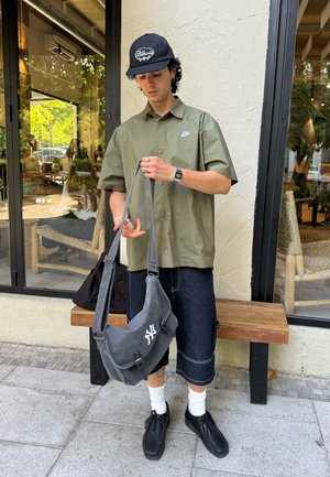 Camisa de manga corta verde oliva, pantalones cortos de mezclilla oscura, zapatos negros y una bolsa gris con un logo blanco, llevada mientras sostiene la correa de la bolsa.