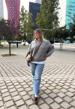 Mujer joven con cabello rubio, vestida con suéter gris, jeans azules, zapatos marrones y bolso cruzado, de pie en una plaza pavimentada con árboles y edificios modernos.