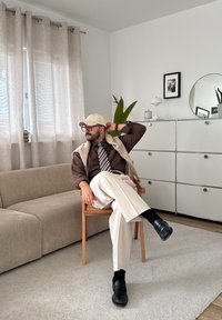 Mężczyzna w beżowej czapce, brązowej koszuli, w paski krawacie i białych spodniach siedzi ze skrzyżowanymi nogami na drewnianym krześle w minimalistycznym salonie.