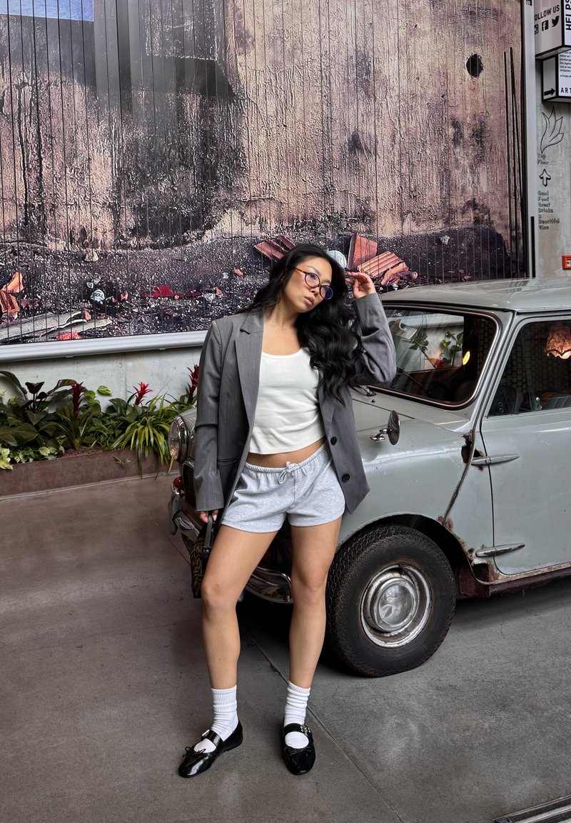 Harmaa bleiseri valkoisen paidan, harmaiden shortsejen ja mustien kenkien kanssa, joissa on valkoiset sukat. Taustalla vintage-auto, vihreitä kasveja ja teksturoitu seinä.