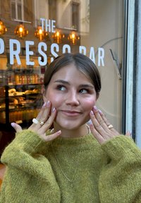Mlada žena u zelenom džemperu pokazuje ružičaste nokte s točkicama i prstenje, smiješi se s kafanskog prozora iza kojeg piše "THE PRESSO BAR."