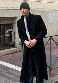 Jeune homme portant un long manteau sombre, un bonnet et un pull, appuyé sur une rambarde métallique dans une rue de la ville aux murs en pierre.