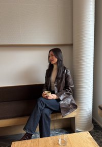 Femme aux cheveux longs et foncés assise sur un banc en bois dans un café, tenant une tasse, portant une veste en cuir marron et un jean foncé.