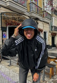 Svart skinnjakke med vide ermer, med hvite Adidas-striper. Blå caps, briller og grå jeans er også synlige. Utendørs setting.
