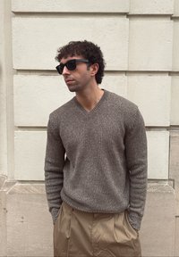 Άνδρας με σγουρά μαλλιά φοράει μαύρα γυαλιά ηλίου, καφέ πλεκτό πουλόβερ και μπεζ παντελόνι, στέκεται μπροστά σε έναν τοίχο από λευκό πέτρινο υλικό.