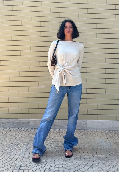 Top blanco con nudos, vaqueros de pierna ancha azules y sandalias negras con acentos dorados. Bolso de estampado de leopardo al hombro; fondo texturizado.