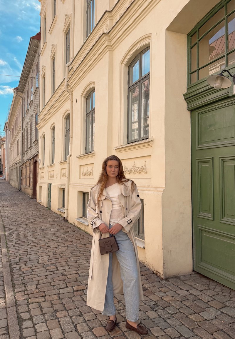 Jeune femme en trench-coat beige et jeans tenant un sac, debout dans une rue pavée près d'un bâtiment crème avec une porte verte et de grandes fenêtres.