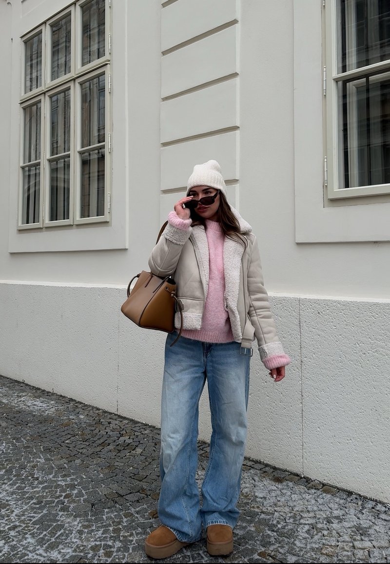 Μπεζ bomber τζάκετ από shearling, ροζ πουλόβερ, oversized ανοιχτό μπλε τζιν, καφέ μποτάκια με πλατφόρμα, λευκό σκουφάκι, καφέ τσάντα χειρός, γυαλιά ηλίου.