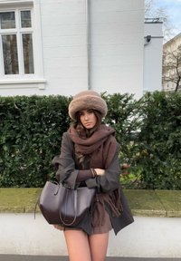 Brūns mākslīgās kažokādas cepure, apvienota ar tumši brūnu šalli un tumši brūnu ādas rokassomu. Tumši svītrains bleizers virs brūnas blūzes.