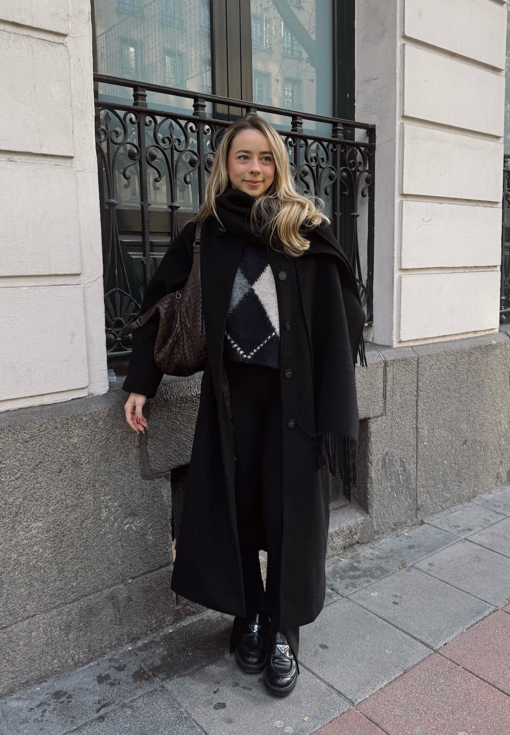 Long manteau noir sur une tenue noire, pull à motifs gris et blancs, sac noir, et chaussures noires brillantes ; debout contre un mur en pierre.