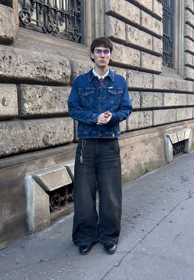 Chaqueta de mezclilla azul con dos bolsillos en el pecho, sobre una camisa blanca y corbata roja. Combinada con unos pantalones vaqueros anchos negros y zapatos negros, de pie contra una pared de piedra.