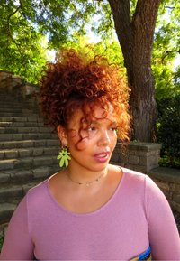 Kvinne med krøllete rødt hår og store grønne blomsterøredobber står utendørs nær steintrappen og et tre, iført en rosa topp og halskjede.