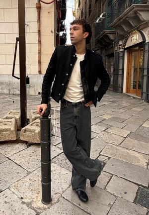 Joven apoyado en un poste negro sobre un pavimento de piedra, vistiendo chaqueta negra, camisa blanca y jeans anchos, de pie en una calle histórica europea.