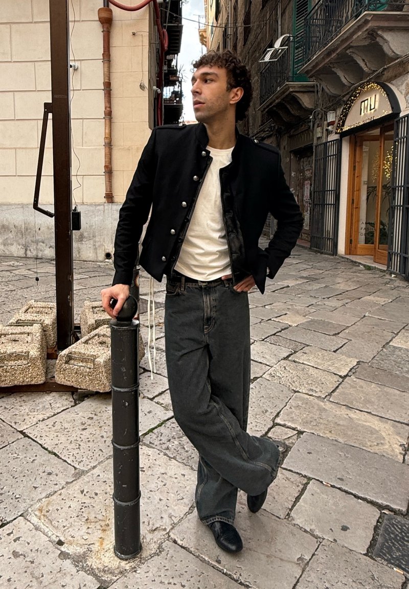 Giovane uomo appoggiato a un palo nero su un pavimento in pietra, indossa una giacca nera, una camicia bianca e jeans larghi, in piedi su una strada storica europea.