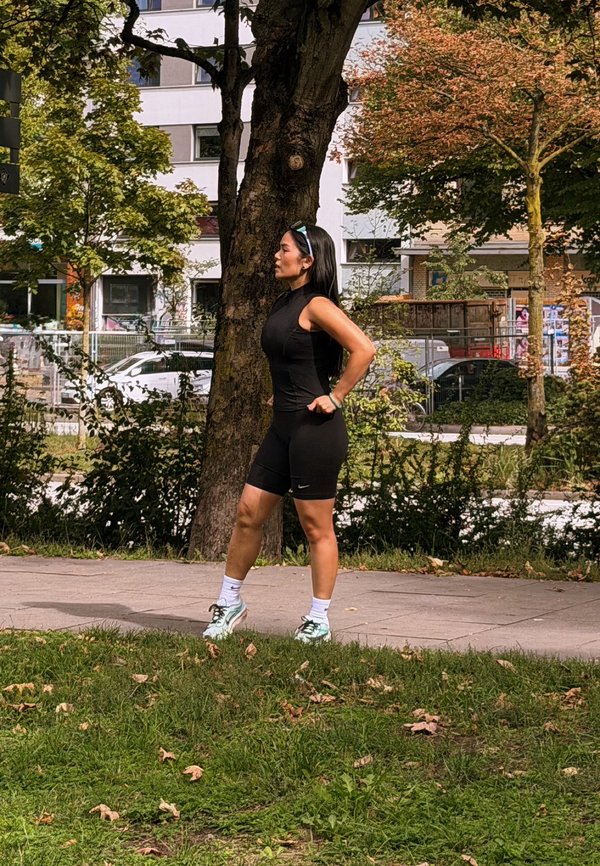 Woman in black athletic outfit stands with hands on hips on a park pathway, surrounded by trees and urban buildings in the background.
