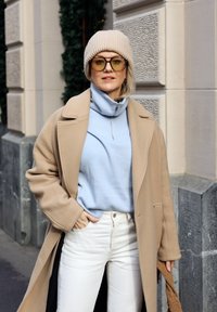 Manteau en laine beige par-dessus un pull bleu clair à col haut, jean crème et bonnet côtelé beige. De grandes lunettes de soleil beiges complètent le look.