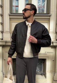 Mann in dunkelbrauner Bomberjacke, cremelangem Strickpullover, grauen Hosen, Sonnenbrille, der eine beigefarbene Tragetasche in einer städtischen Umgebung vor einer Gebäude-Fassade hält.