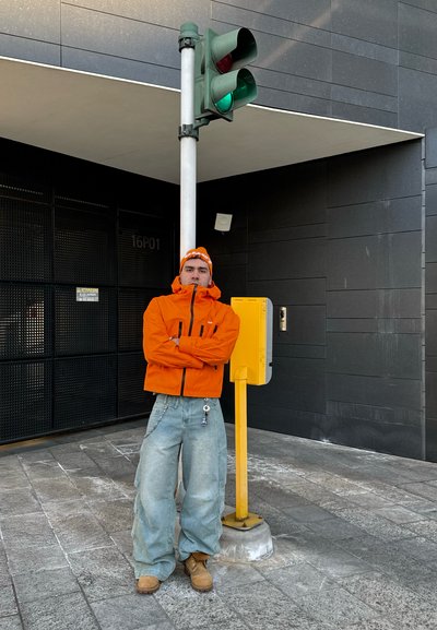 Chaqueta naranja con cremallera, jeans holgados de color azul claro, botas marrones; de pie cerca de un semáforo verde y una señal de peatones amarilla en un entorno urbano.