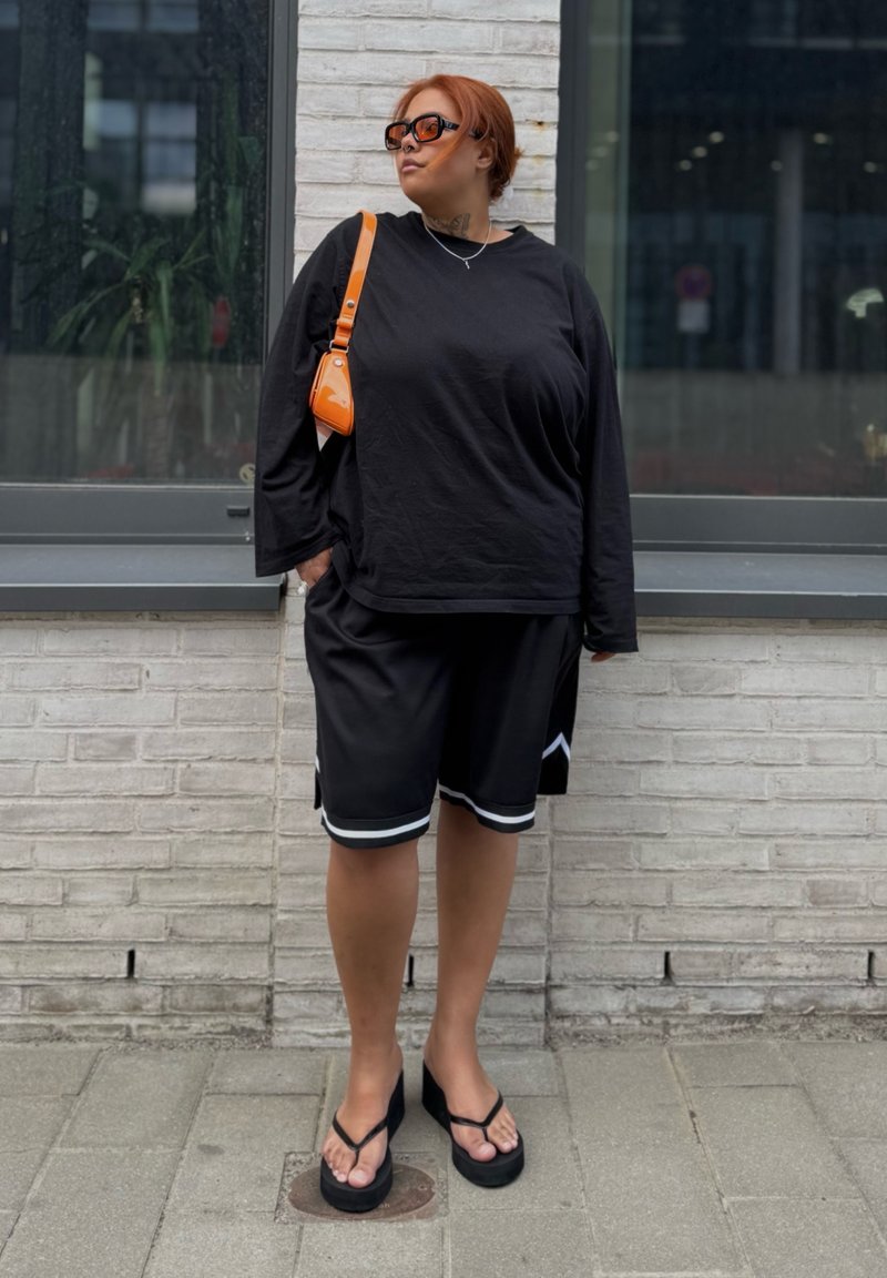 Person mit roten Haaren, die ein schwarzes Langarmshirt, schwarze Shorts, eine orangefarbene Umhängetasche, Sonnenbrille und schwarze Keilsandalen trägt und vor einer weißen Backsteinmauer steht.