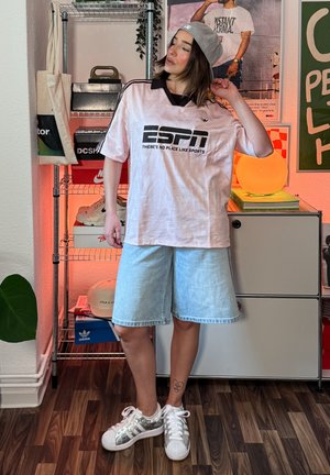 Joven que lleva una camiseta deportiva color rosa claro de ESPN, gorra gris al revés, pantalones cortos de mezclilla y zapatillas blancas, de pie en una habitación decorada.