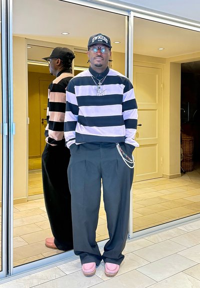 Hombre vestido con camisa a rayas blancas y negras, pantalones negros con cadena, zapatos rosas y gorra negra, de pie en el interior junto a puertas con espejos.