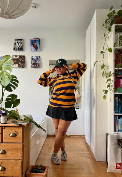 Camisa de manga larga a rayas en azul marino y mostaza, falda negra, zapatillas de cuadros, gorra verde y un pequeño bolso cruzado en un entorno interior luminoso.