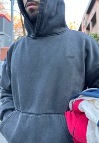 Homme portant un sweat à capuche sombre, debout en plein air, tenant une veste pliée rouge, blanche et bleue devant un arrière-plan urbain.